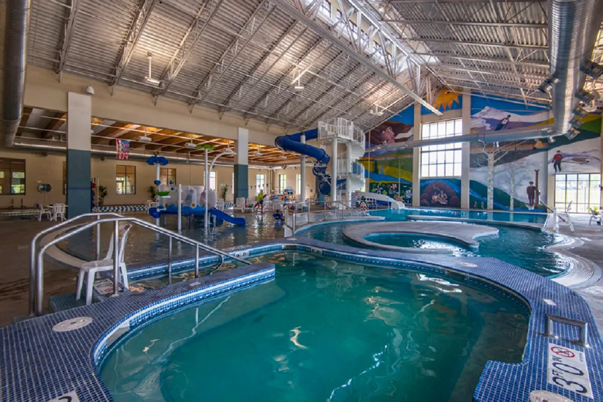 A brightly lit indoor recreation center scene features children gleefully playing on colorful water slides, surrounded by lively wall murals. The spa area and spacious pools provide a vibrant and energetic space perfect for families with kids.