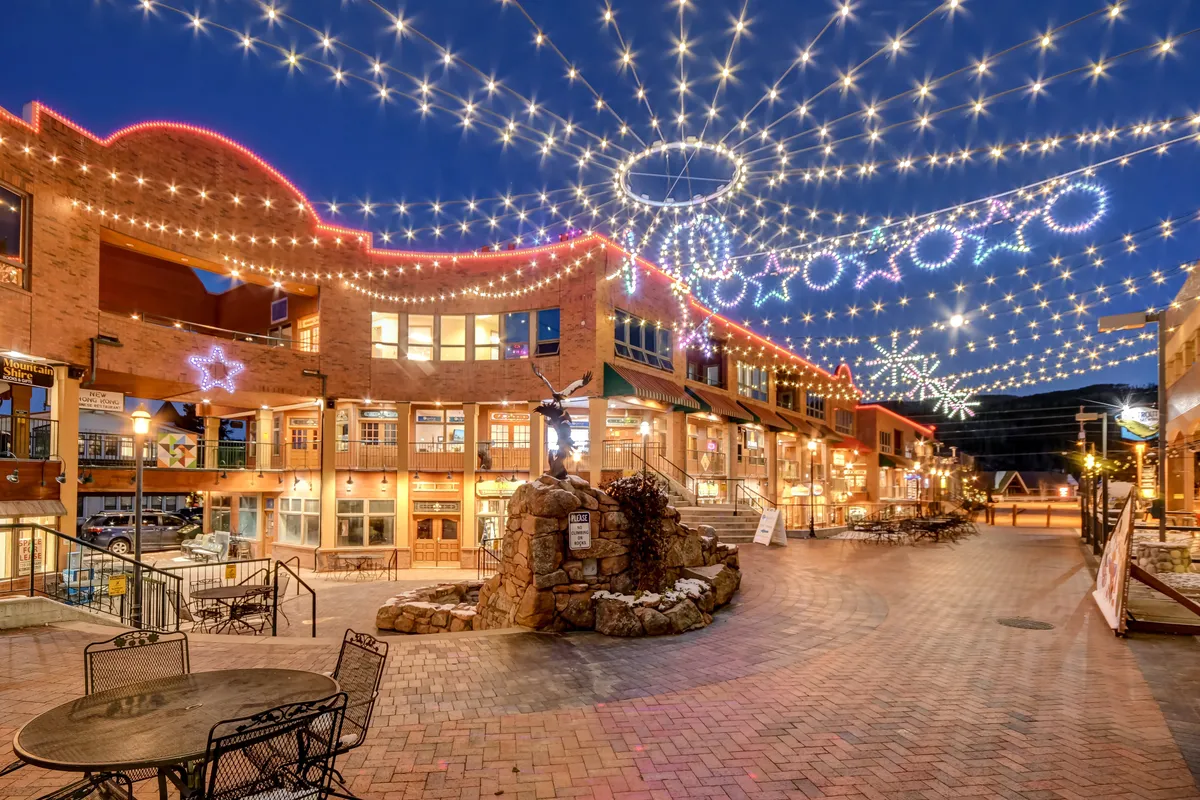 A charming brick pathway lined with boutique shop windows, illuminated by string lights above. In the center is a stone fountain, and the scene is lively with people dining, shopping, and socializing in a festive, walkable environment.