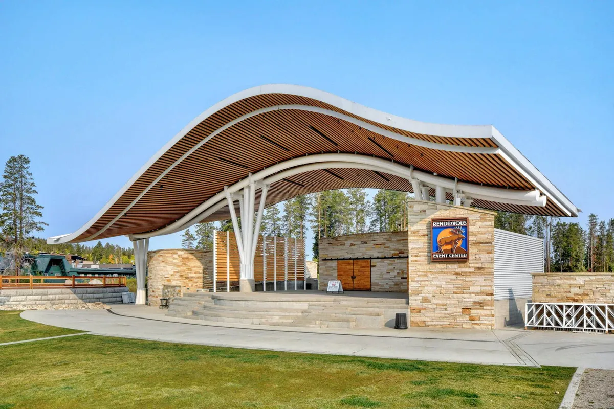 An exterior shot featuring the event center with its distinctive curved roof and stone facade. The area is alive with signage promoting community events, concerts, and outdoor gatherings, presenting a lively and welcoming environment.