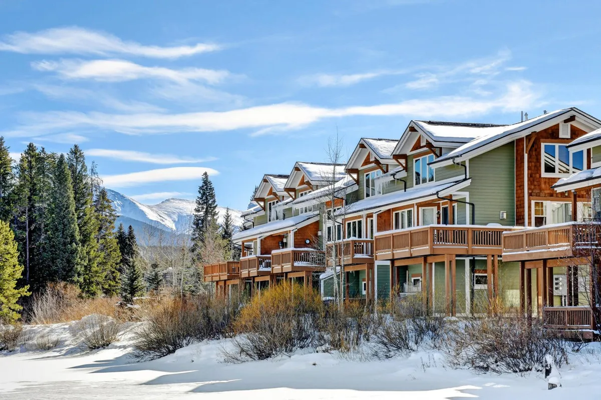 The image shows a striking multi-story villa nestled amid a pristine, snow-covered landscape, flanked by tall evergreens and with breathtaking views of distant, rugged mountains. The tranquil scene evokes a sense of cozy seclusion and adventure, inviting guests to imagine winter activities right from their doorstep.