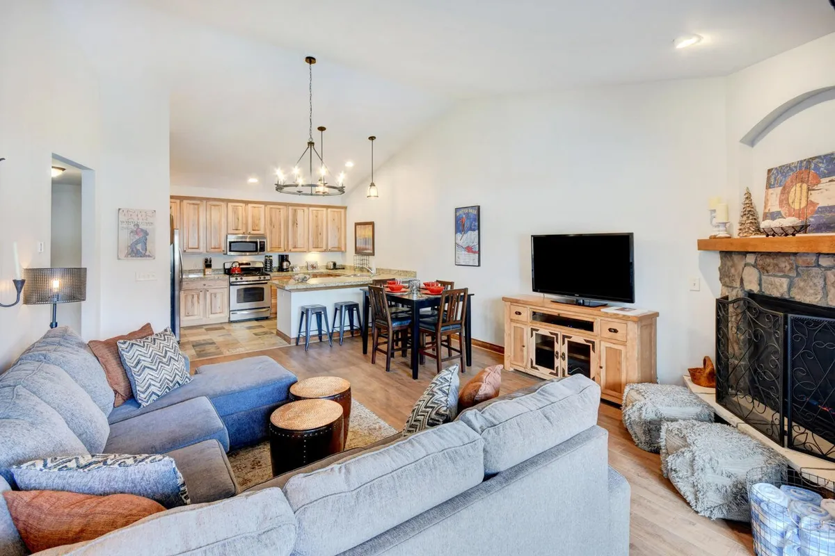 A warm and inviting great room where the kitchen island and a dining table share the space with a large sectional sofa and a crackling fireplace. The scene captures the multi-functional heart of the home, where meals, gatherings, and relaxation blend seamlessly.