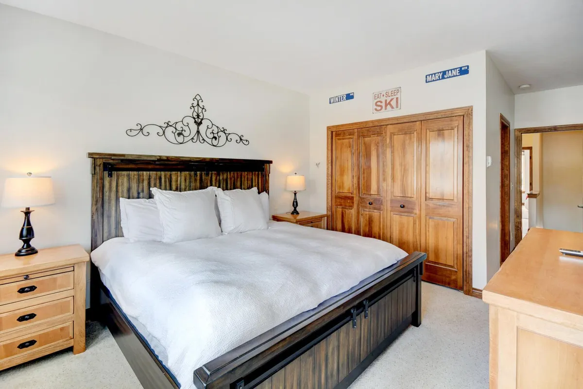 A peaceful bedroom with a king-size bed dressed in crisp white bedding, complemented by wooden nightstands and closet doors. Soft natural light streams in from a window, highlighting decorative wall accents and creating a serene, comfortable atmosphere.