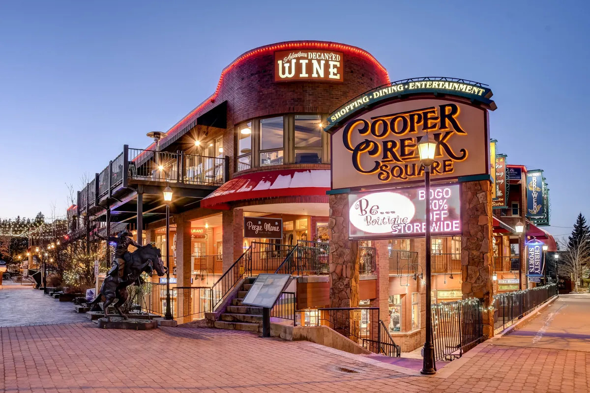 The Cooper Creek Square entrance is visible with its prominent sign, decorative lighting, and a welcoming wine shop entrance. The area buzzes with activity, hinting at shopping, dining, and live entertainment opportunities in the heart of downtown.