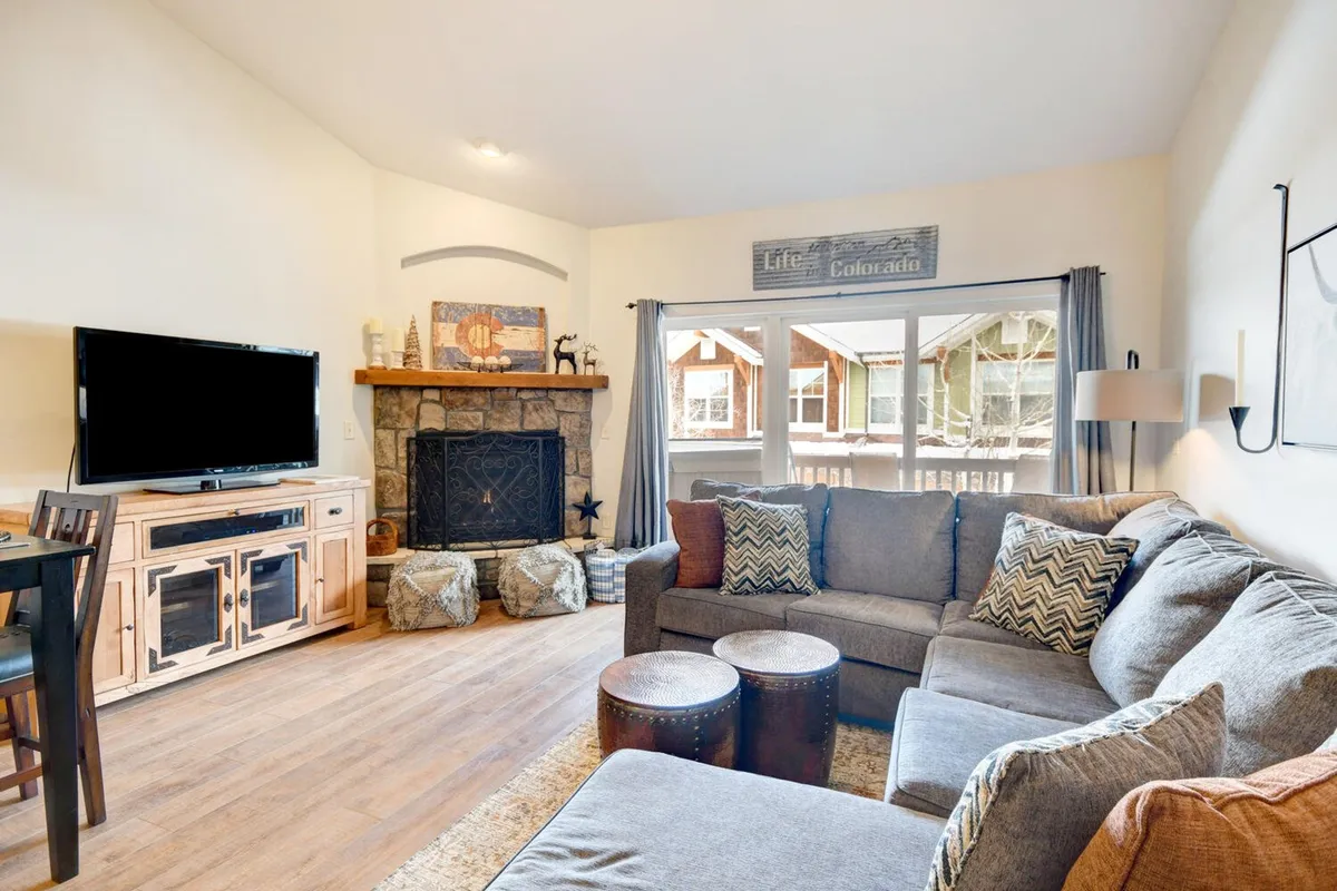 A cozy living room featuring a plush sofa, a modern fireplace, a sleek coffee table, and a wall-mounted television. Sunlight streams through expansive windows, illuminating the gathering space and offering glimpses of the mountains beyond.